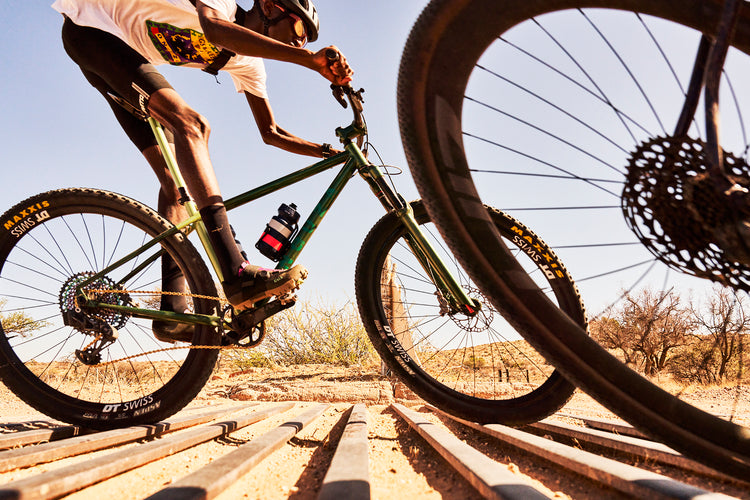 A black Namibian mountain biker riding an ONGUZA Rooster hardtail mountain bike in the Boomslang Green color, crosses a cattle grate. He stands up off his saddle, legs extended to soften the rumbling metal crossing. In the foreground another cyclists' wheel is distorted, moving out of the image. They are in the Namibian desert between Windhoek and Spreetshoogte Pass. Photo by Ben Ingham