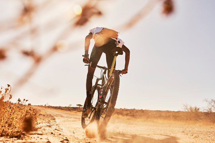 A black Namibian mountain bike rider accelerates on a dusty gravel road in the Namibian desert. His body is slightly silhouetted against a pale blue sky. Dust trails behind his bike as he climbs out of his saddle to accelerate. His form is slightly obscured by a thorn branch, out of focus in the foreground. He is riding an ONGUZA Rooster hardtail mountain bike in the Boomsland Green color. Photo by Ben Ingham.