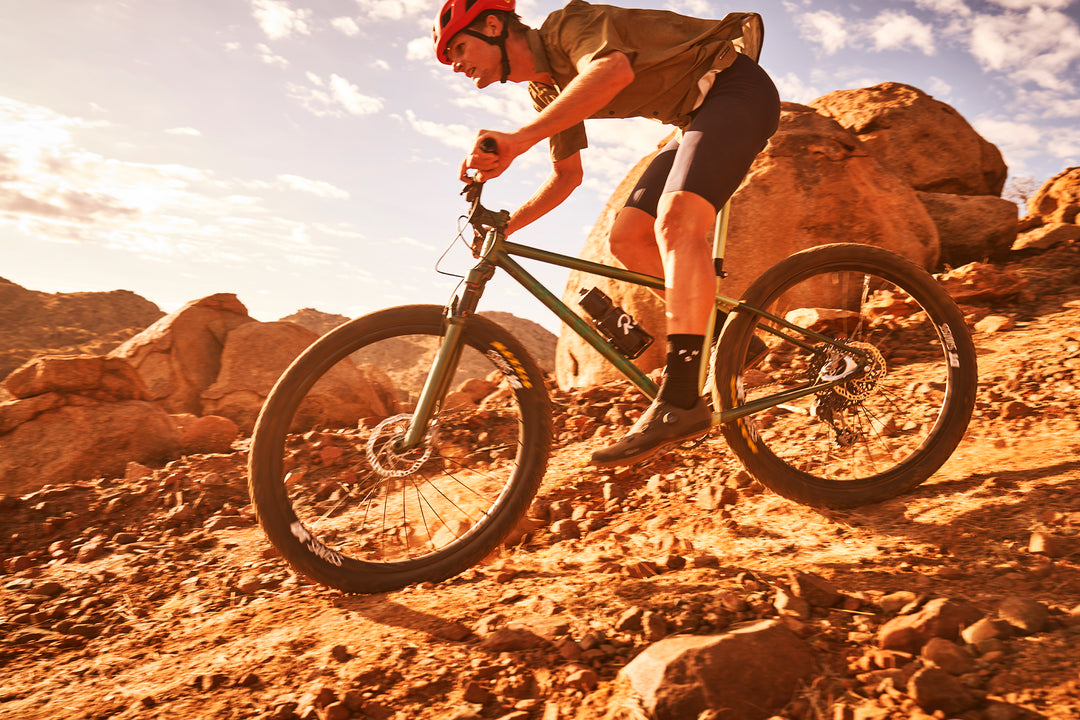 A caucasian Namibian man rides an ONGUZA Rooster hardtail mountain bike in Boomslang Green down a rocky path. He is standing up hovering above his saddle, eyes fixed and concentrated on the path ahead. He is surrounded by oversized, prehistoric boulders, glowing orange in the early morning sun. He is wearing a red POC helmet, a loose CIOVITA shirt and black bib shorts. Dust kicks up underneath his tyres. Photo by Ben Ingham