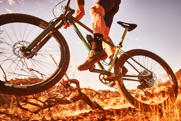 A caucasian Namibian mountain bike rider jumps over a dead tree in the rocky Namibian desert, lifting his front wheel into the air. His fizik cycling shoes are clipped in and as he lifts off, his toes peak downward, heels in the air. He is riding a Boomslang Green ONGUZA Rooster hardtail MTB. The sky is pale blue and the earth below is a dusty orange. Photo by Ben Ingham.