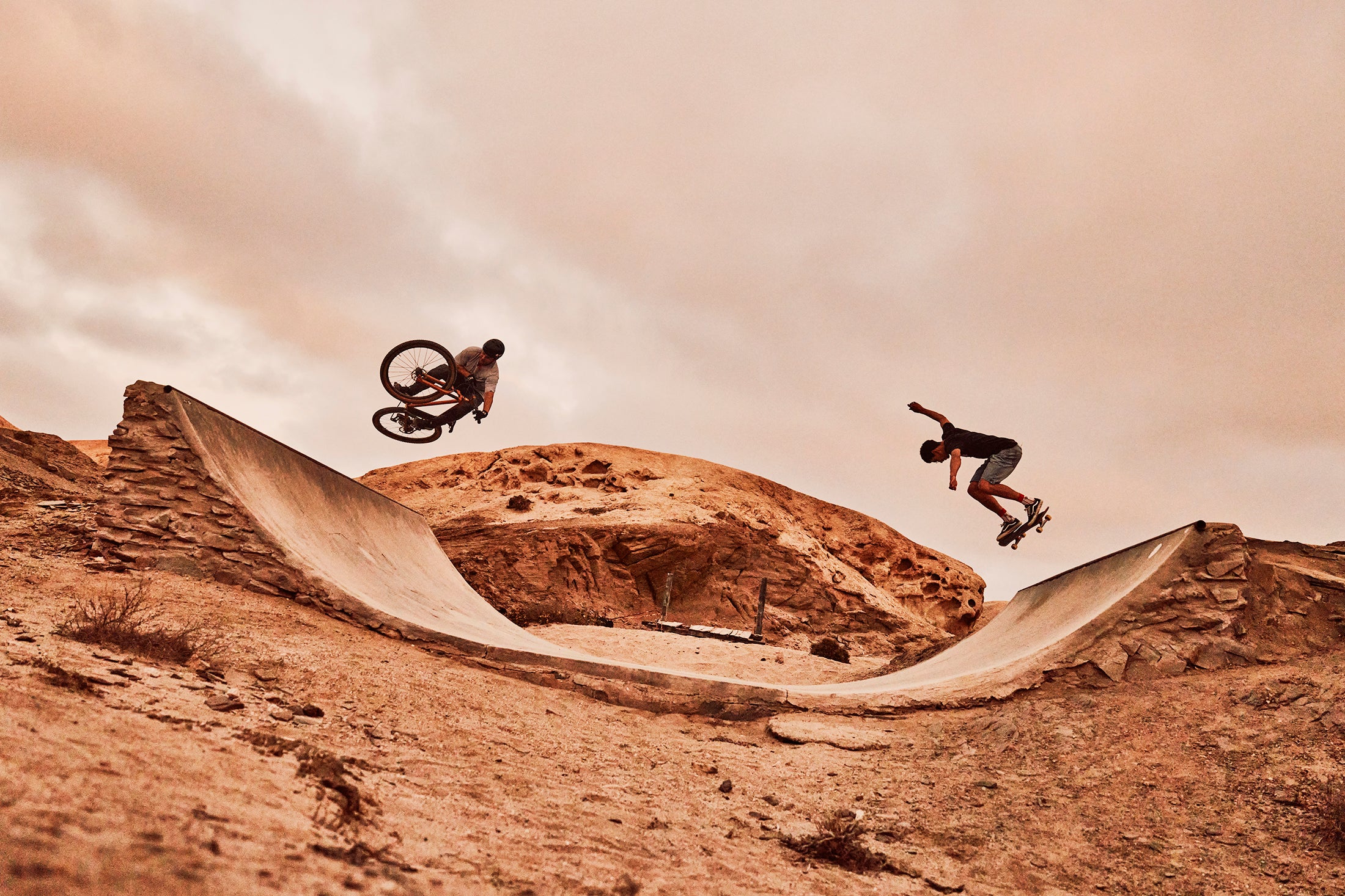 A BMX rider and skateboarder take to the air on a homemade half-pipe just outside of Swakopmund, Namibia. They are outlined against the grey cloudy coastal sky. The BMX rider is riding an ONGUZA Rooster hardtail mountain bike, with the bike horizontal in the air nearly a meter off the surface. The skateboarder throws his arms up in balance. The desert around them is dry and salt-encrusted. Photo by Ben Ingham