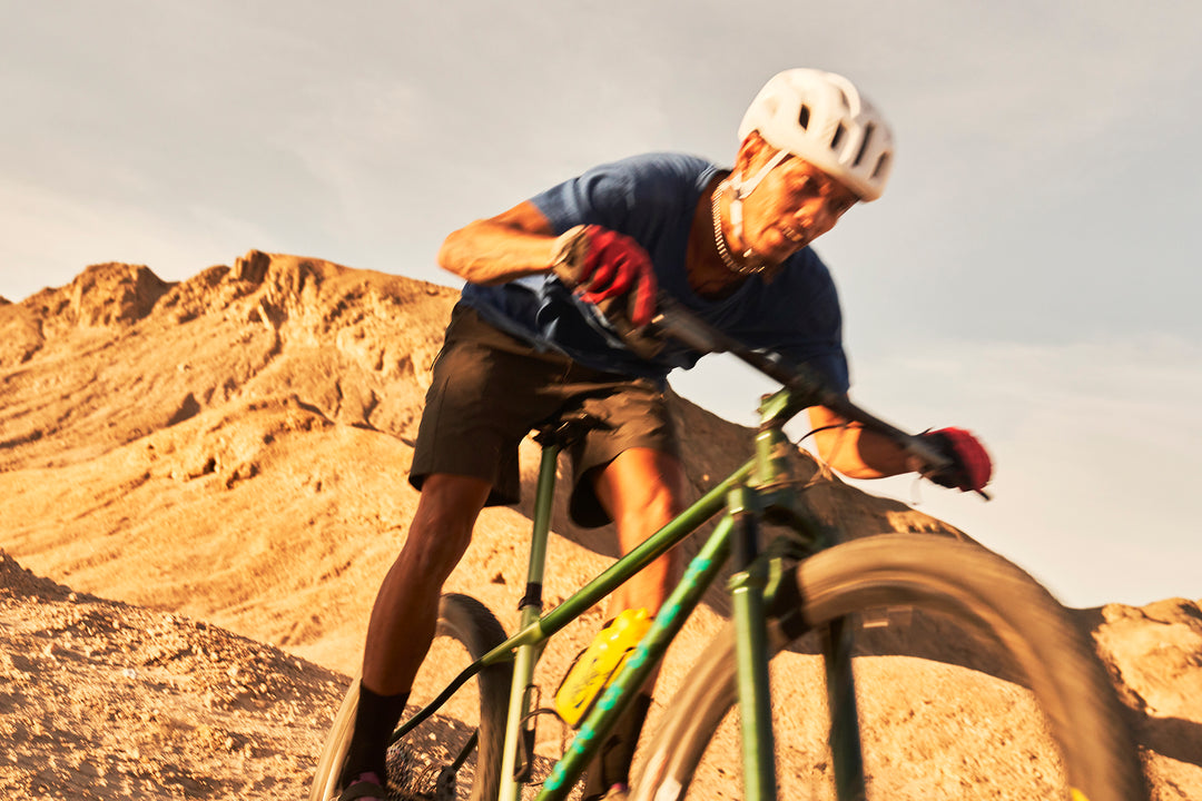 A Namibian man of color descends a loose, rocky single track trail on an ONGUZA Rooster hardtail mountain bike in the Boomslang Green color. He is riding fast and his face is concentrated, mouth open, eyes fixed on the path ahead. He is wearing a white POC helmet, red gloves, loose black shorts and a simple, loose blue t-shirt. Photo by Ben Ingham