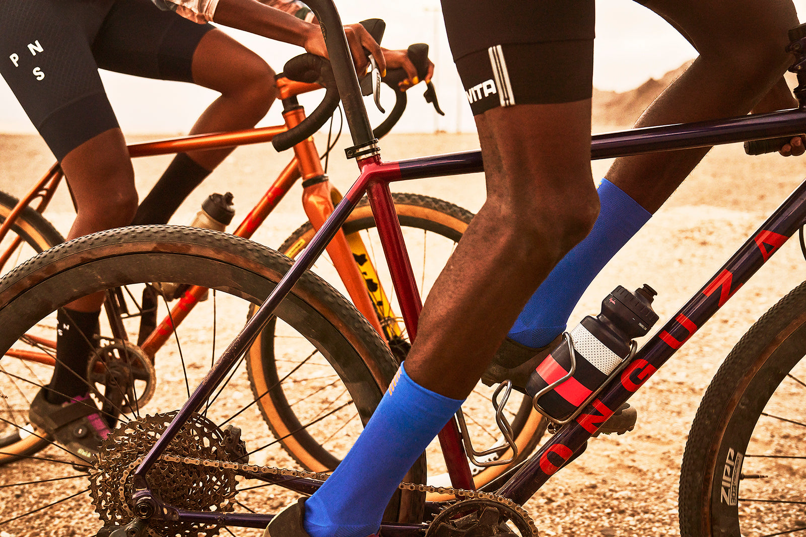 Two black Namibian cyclists, a man in the foreground and a woman in the background, riding in the Namibian desert on ONGUZA Goat Gravel bikes. The shot is cropped around the bikes and their legs. The bike in the foreground is Eastwind Purple, and the background is Gold Dune colors. The man is wearing electric blue socks which contrasts with the colors of the bikes. Photo by Ben Ingham