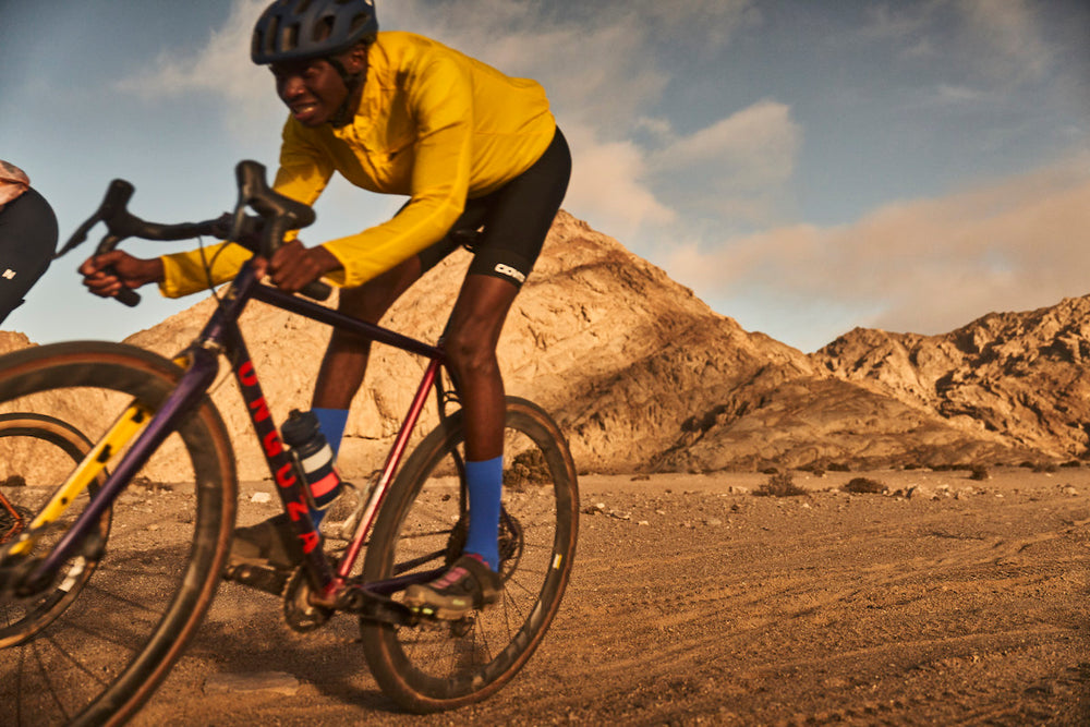 A black Namibian cyclist races through the mountainous desert by Rossing Mountain outside of Swakopmund, Namibia. He is hovering off his saddle, eyes fixed on the path ahead. He is riding an Eastwind Purple color Goat Gravel bike with tan sidewall tyres. His blue socks and yellow Patagonia windbreaker pop in the pale, dusty landscape. Photo by Ben Ingham