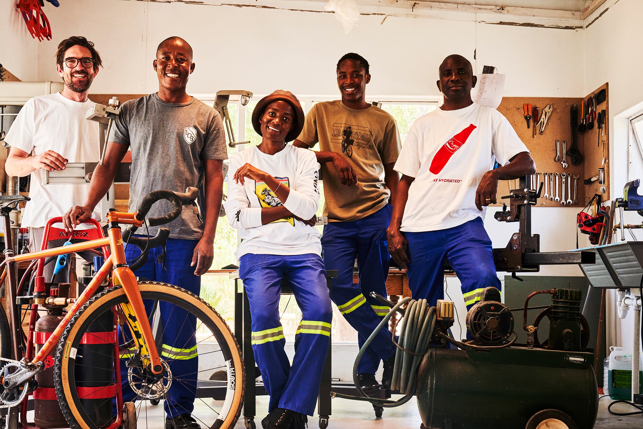 Dan Craven, Petrus Mufenge, Tilomwene Mundjele, Sakeus Mufenge and Sakaria Nkolo, the builders of ONGUZA bicycles, stand and lean against a table in the ONGUZA factory in Omaruru, Namibia. They are relaxing and smiling at the camera, evoking pride. In the foreground is an ONGUZA Gold Dune Goat Gravel bike with it's golden shade glowing in the sunlight coming in through a window to the left. They are surrounded by their builders' tools and equipment including an air compressor, brazing-welding kit and jig.