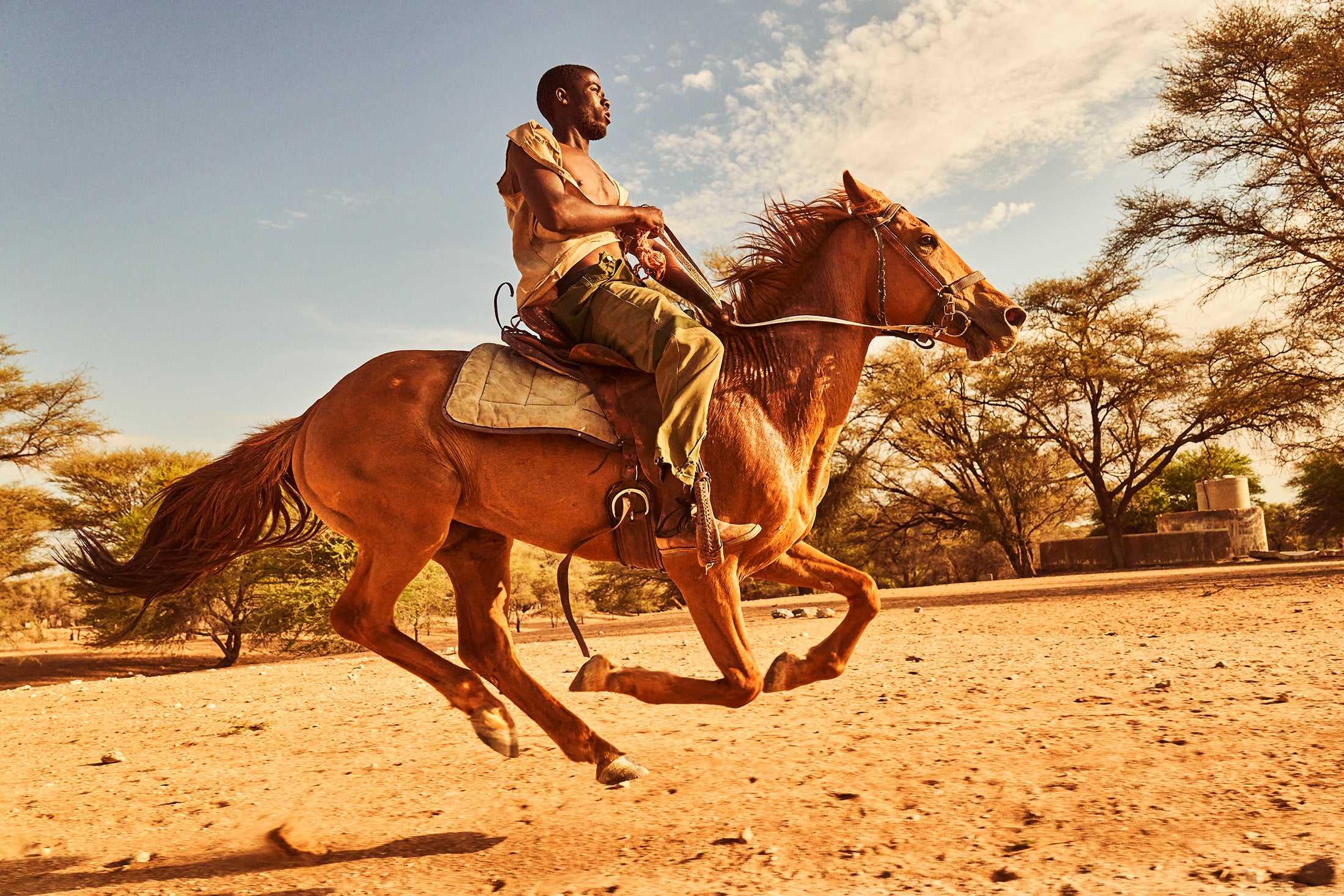 A black Namibian man rides a golden colored horse in the dried riverbed just outside Okambahe, Namibia. He looks up at the horizon in front of him. His horse is galloping with all hooves off the ground. The man is wearing a simple khaki pant and loose shirt, open and exposing his chest to the late afternoon sun. The horse is saddled with a simple leather saddle and blanket, soaked with sweat and dirt. Photo by Ben Ingham