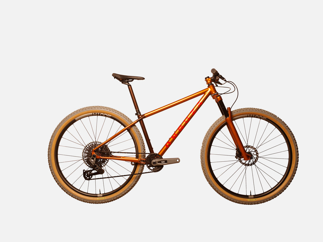 A side-on view of the ONGUZA Rooster hard tail mountain bike in a size small, in the Gold Dune color. The bike features a suspension fork, also painting in the golden color. The seat tube and seat post are a chocolate brown with the front and rear triangles in a golden bronze color that glows in the sunlight. The ONGUZA wordmark on the down tube is in a bright fire red color. The carbon wheels on the bike are made by South Industries in South Africa and feature grey-tread tyres with tan side-walls.