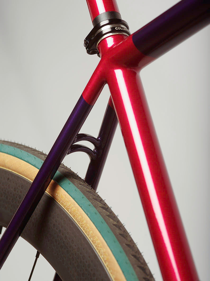 The rear double bridge and seat tube of an ONGUZA road bike in berry pink and purple. Highlights the fine brazing and filing workmanship. Attached is a ZIPP deepsection wheel with Ultradynamico tyres in an ultramarine green and tan sidewalls. 