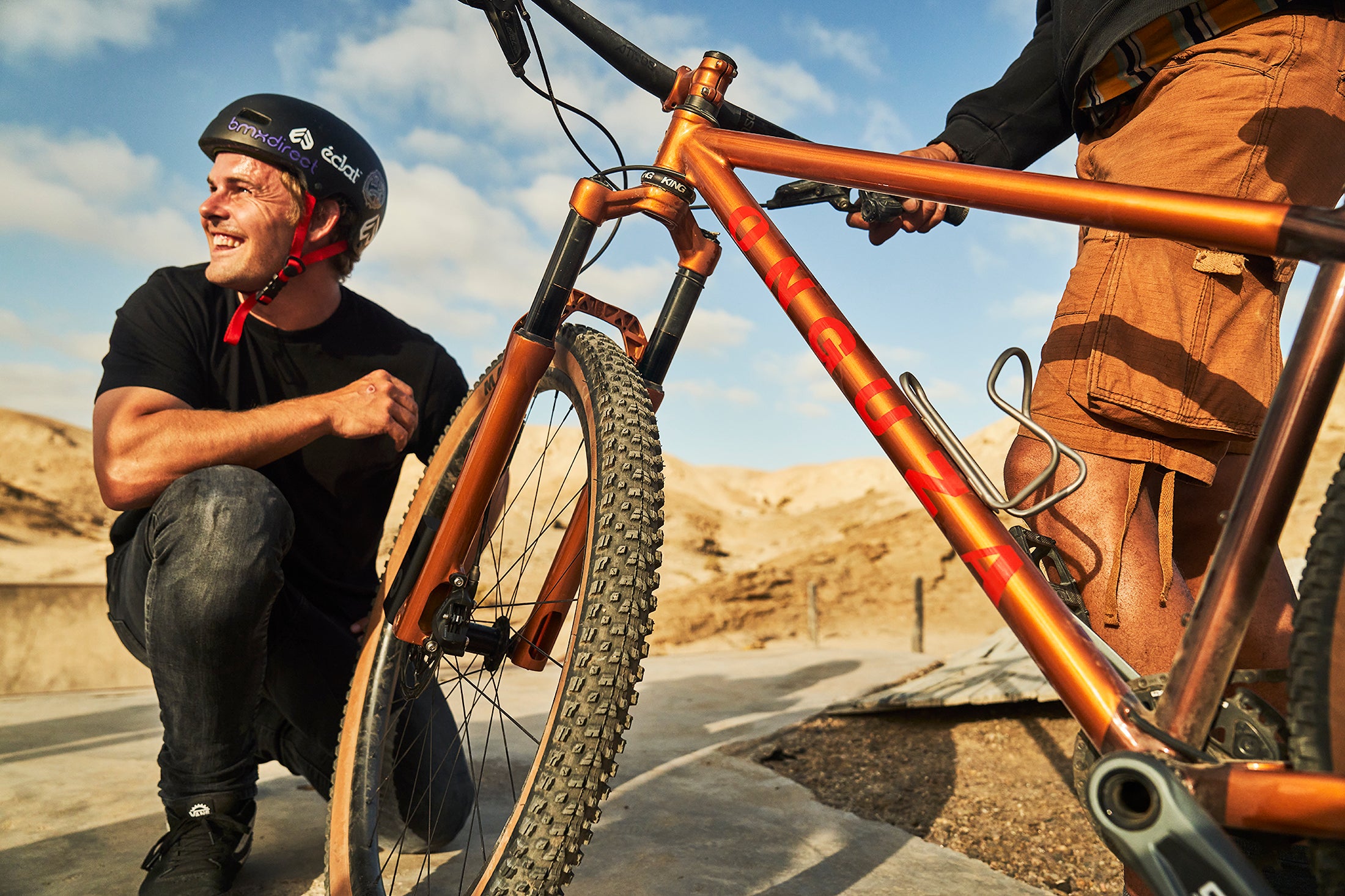 A caucasian Namibian BMX rider kneels next to an ONGUZA Rooster hardtail mountain bike in Gold Dune color. He is helmeted and smiling to someone off-camera. Standing next to him with the bike is a Namibian man of color, wearing cargo shorts. His body is cropped out of the image. They are standing at a half-pipe in the desert outside Swakopmund Namibia. Photo by Ben Ingham