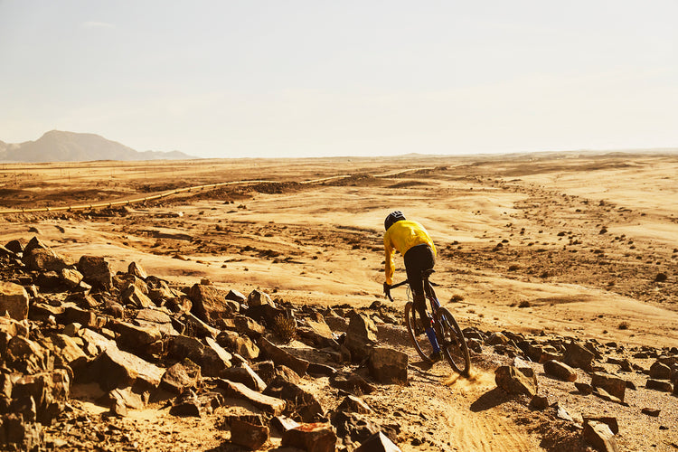 A black Namibian man rides an ONGUZA Goat Gravel bike on a technical piece of single track in the desert just outside of Swakopmund, Namibia. Rossing Mountain looms in the background. The waterpipe which pumps water from inland to the coast snakes past on the ground below the trail. The rider is wearing an old yellow Patagonia wind jacket, a black POC helmet, and black CIOVITA cycling bib shorts. Photo by Ben Ingham.